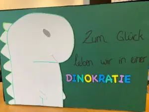 Wahlurne des Jugendzentrums Funzel in Krefeld zur U16-Bundestagswahl 2025. Auf grünem Tonkarton ist links ein grauer Dino aufgeklebt, der von der Seite gezeigt wird. Rechts daneben steht in schwarzer Schreibschrift "Zum Glück leben wir in einer". Darunter steht in einzelnen Großbuchstaben aus Tonkarton in lila, blau, türkis und gelb das Wort "Dinokratie".