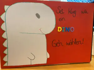 Wahlurne des Jugendzentrums Funzel in Krefeld zur U16-Bundestagswahl 2025. Auf rotem Tonkarton ist links ein grauer Dino aufgeklebt, der von der Seite gezeigt wird. Rechts daneben steht in schwarzer Schreibschrift "Sei klug wie ein". Darunter steht in einzelnen Großbuchstaben aus Tonkarton in gelb, grün, blau und lila das Wort "Dino". Darunter ist wieder in schwarzer Schreibschrift zu lesen "Geh wählen!"
