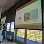 Großer Bildschirm mit Präsentation des PerspektivWechsel-Workshops auf dem DJHT 2025. Links sind zwei Referent_innen am Stehpult zu sehen.