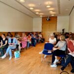 Ein Seminarraum mit rechts und links Stuhlreihen auf denen Teilnehmende sitzen und zuhören. in der Mitte ist ein freier Gang.