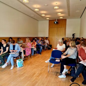 Ein Seminarraum mit rechts und links Stuhlreihen auf denen Teilnehmende sitzen und zuhören. in der Mitte ist ein freier Gang.