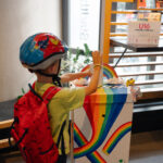 Kind mit buntem Fahrradhelm wirft einen Wahlzettel in eine regenbogenfarbene Wahlurne